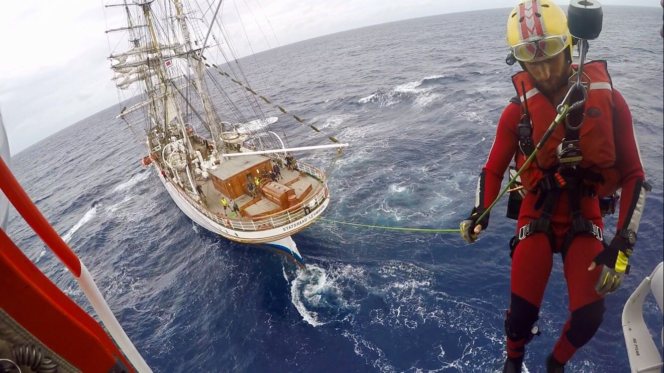 Rescatador descendiendo de un helicóptero y, al fondo, barco de época